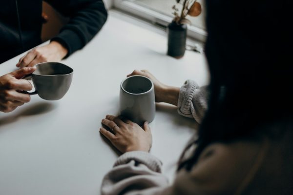 Two people holding coffee mugs across a table during a friendly conversation